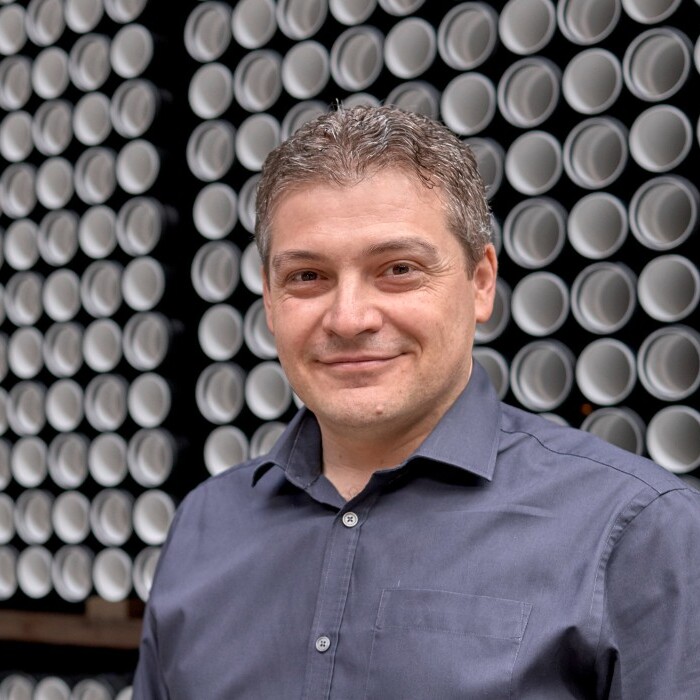 A man stands smiling in front of a wall full of pipes.