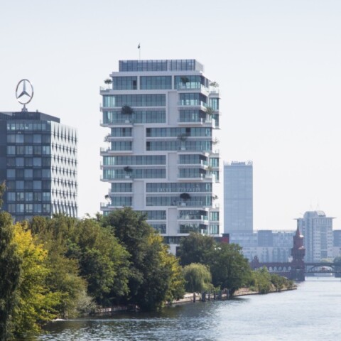 Moderne Gebäude am Ufer eines Flusses, umgeben von Bäumen und einer Stadtlandschaft im Hintergrund.