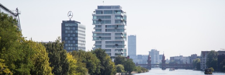 Moderne Gebäude am Ufer eines Flusses, umgeben von Bäumen und einer Stadtlandschaft im Hintergrund.