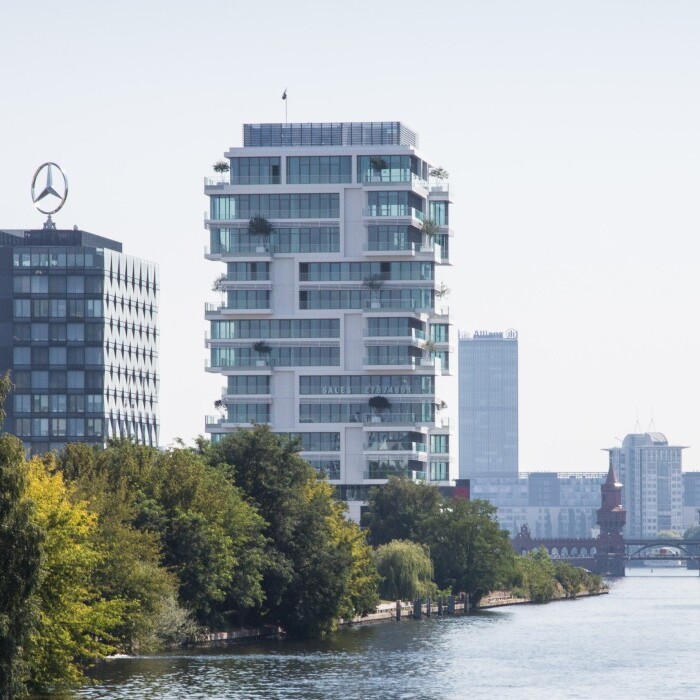 Moderne Gebäude am Ufer eines Flusses, umgeben von Bäumen und einer Stadtlandschaft im Hintergrund.