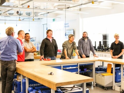 Eine Gruppe von sieben Personen, steht an einem langen Tisch in einem gut beleuchteten Workshopraum.