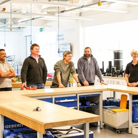 Eine Gruppe von sieben Personen, steht an einem langen Tisch in einem gut beleuchteten Workshopraum.