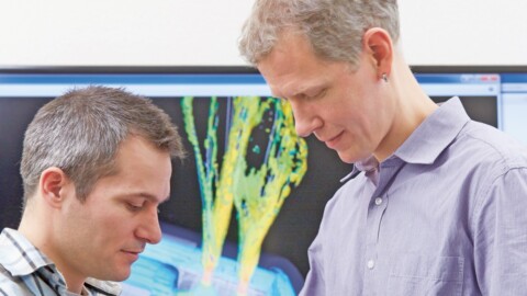 Zwei Männer stehen im Vordergrund und im Hintergrund ist ein Bildschirm mit einer technischen Grafik.