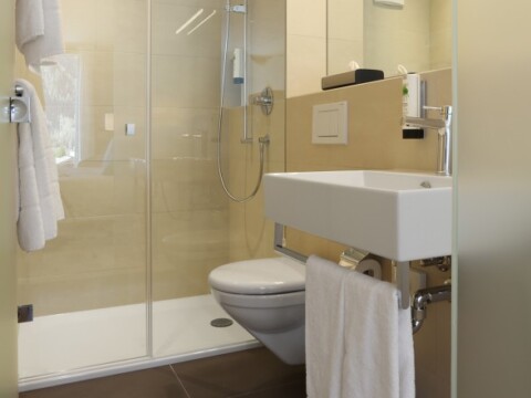 Modern bathroom with a glass shower, toilet, and a wall-mounted sink, featuring beige tiles and a towel hanging.