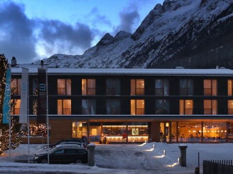 A modern hotel building with large windows, surrounded by snow and mountains, illuminated during dusk.
