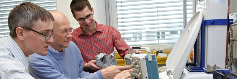 Three men are doing a test in a laboratory setting, discussing components.