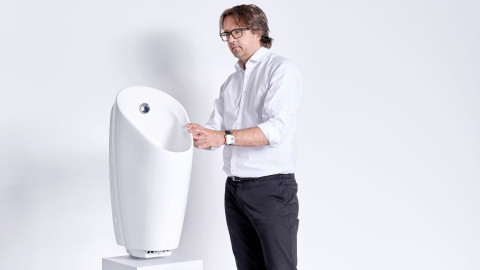 A man in a white shirt is standing next to a modern, white urinal displayed on a pedestal.