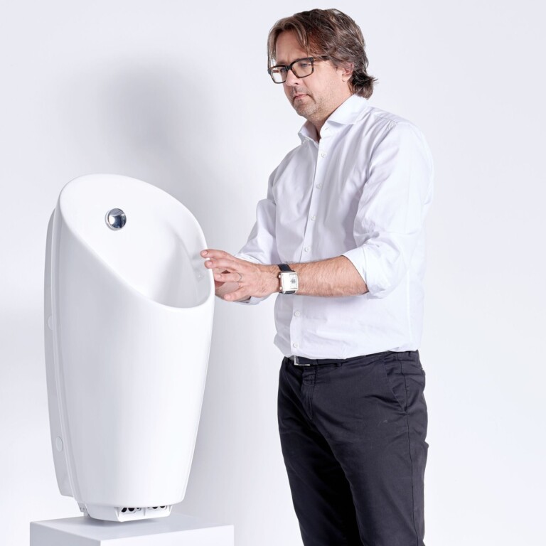 A man in a white shirt is standing next to a modern, white urinal displayed on a pedestal.