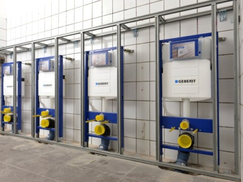 Row of white toilet cisterns mounted on metal frames with plumbing fixtures in a tiled restroom.