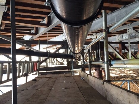 A long black pipe runs horizontally through a framework of metal supports and wooden planks underneath a structure.