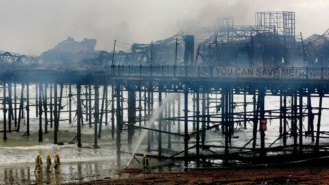 A fire-damaged pier with smoke rising, firefighters in yellow suits working near the water.