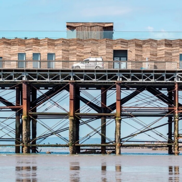 A wooden pier with a modern building on top, supported by metal pillars over a calm sea.