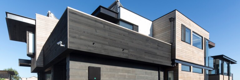 Modern house with a combination of dark wood and light-colored siding, large windows, and a minimalist design against a clear blue sky.
