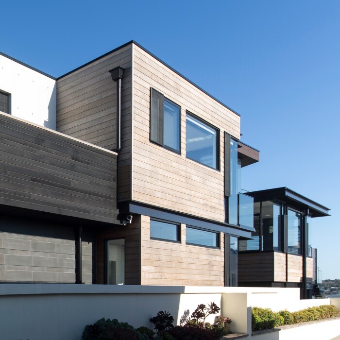Modern house with a combination of dark wood and light-colored siding, large windows, and a minimalist design against a clear blue sky.