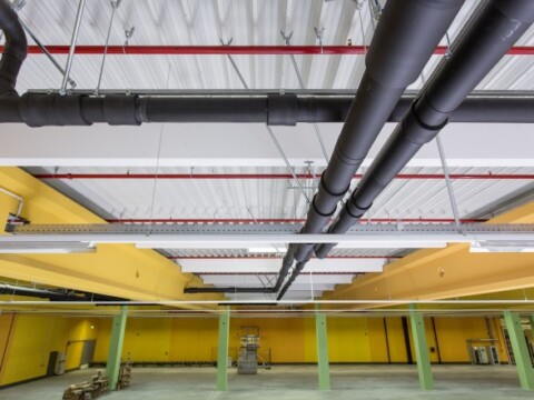 Industrial space with exposed black pipes and yellow walls, featuring an empty floor and green columns.