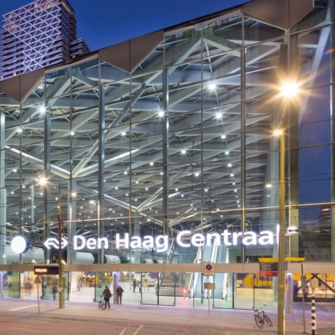 Modern train station with glass facade, illuminated signage reading "Den Haag Centraal," and urban surroundings at dusk.