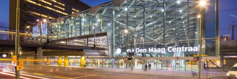 Modern train station with glass facade, illuminated signage reading "Den Haag Centraal," and urban surroundings at dusk.