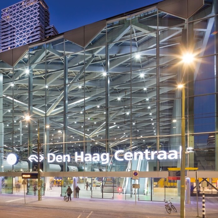 Modern train station with glass facade, illuminated signage reading "Den Haag Centraal," and urban surroundings at dusk.