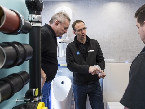 Three men discussing a product in a modern workspace, with pipes and an installation in the background.