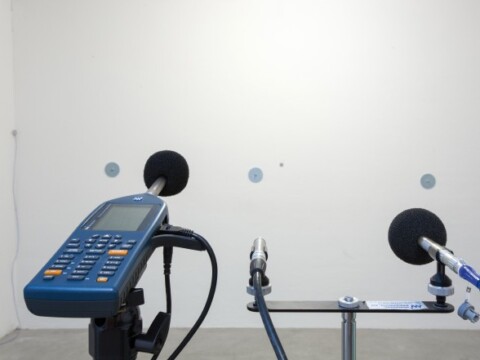 Ein Messgerät mit Mikrofonen, das auf eine weisse Wand ausgerichtet ist, um Schallmessungen durchzuführen.