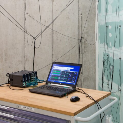 Ein Schreibtisch mit einem Laptop und einem Messgerät in einem Raum mit Betonwänden, an denen mehrere Kabel verlaufen.