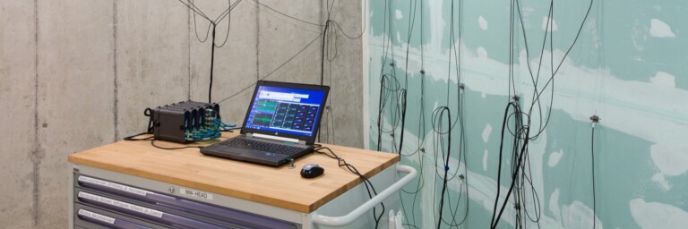 Ein Schreibtisch mit einem Laptop und einem Messgerät in einem Raum mit Betonwänden, an denen mehrere Kabel verlaufen.