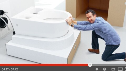 A man crouches beside a large, modern toilet, appearing to push or move it, with a minimalist background and cardboard box in view.