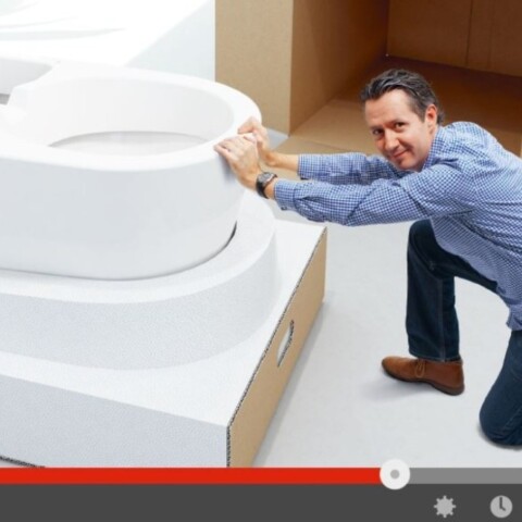 A man crouches beside a large, modern toilet, appearing to push or move it, with a minimalist background and cardboard box in view.
