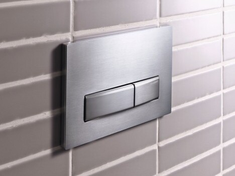 A metallic dual flush toilet actuator plate mounted on a tiled wall.