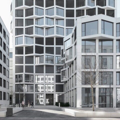 Moderne Architektur mit mehreren Gebäuden in verschiedenen Formen und Fenstergestaltungen, vor einem klaren Himmel.