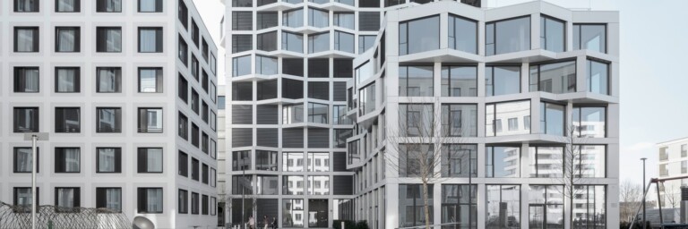 Moderne Architektur mit mehreren Gebäuden in verschiedenen Formen und Fenstergestaltungen, vor einem klaren Himmel.