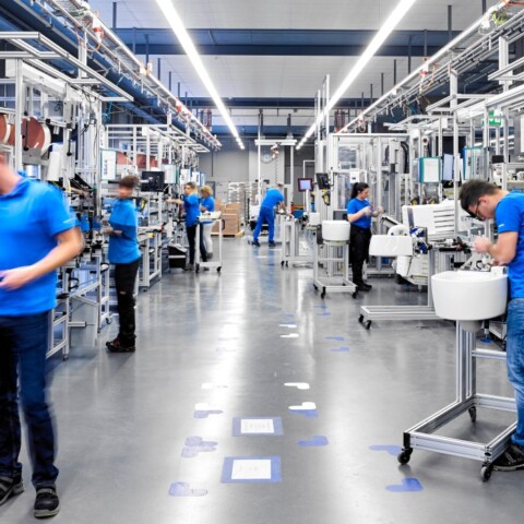 Mehrere Arbeiter in blauen T-Shirts an verschiedenen Produktionsstationen in einer modernen Fabrikhalle.