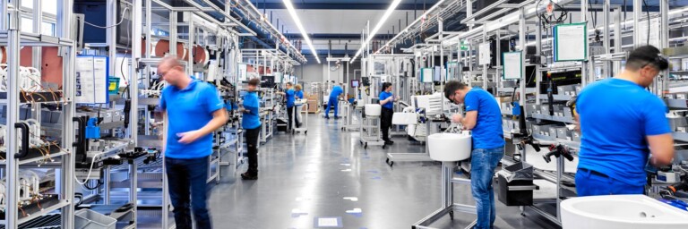 Mehrere Arbeiter in blauen T-Shirts an verschiedenen Produktionsstationen in einer modernen Fabrikhalle.