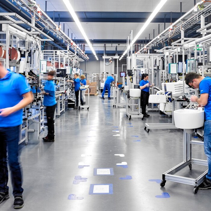 Mehrere Arbeiter in blauen T-Shirts an verschiedenen Produktionsstationen in einer modernen Fabrikhalle.