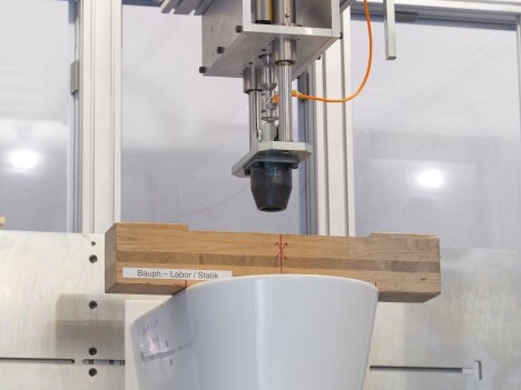 Ein Roboterarm, der über einer weissen Keramikschüssel positioniert ist und auf der ein Holzbrett liegt.