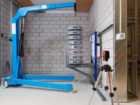 Eine blaue Hebebühne mit gestapelten Gewichten steht in einem Raum mit grauen Betonwänden, daneben ein Vermessungsgerät auf einem Stativ.