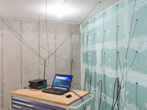 Ein Raum mit Betonwänden, in dem ein Tisch mit Laptop und Geräten steht, umgeben von Kabeln und Sensoren an einer Wand.