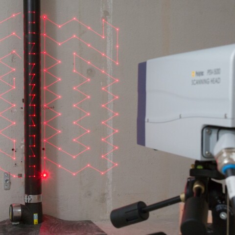 Ein Lasermessgerät auf einem Stativ projiziert ein rotes Muster an eine Wand an ein vertikales Rohr.