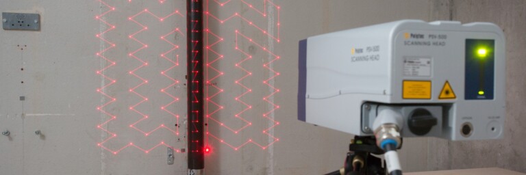 Ein Lasermessgerät auf einem Stativ projiziert ein rotes Muster an eine Wand an ein vertikales Rohr.