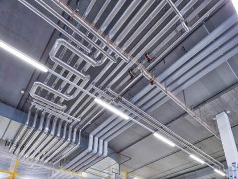 Ein Blick auf die Decke eines industriellen Raums mit zahlreichen sichtbaren Rohrleitungen und Beleuchtung.