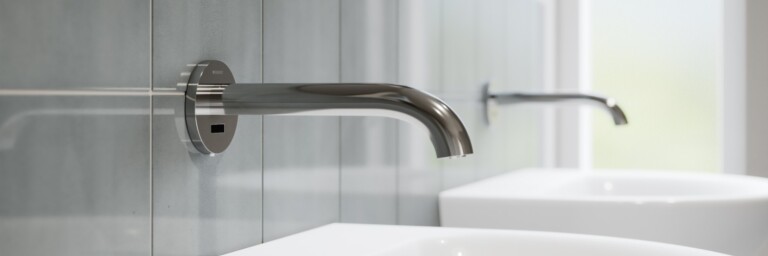 Modern bathroom scene featuring two sleek silver faucets mounted on the wall over white sinks against a light grey backdrop.