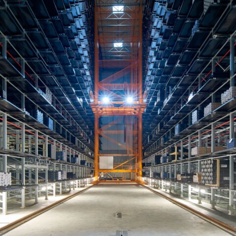 Ein grosser, gut beleuchteter Lagerraum mit hohen Regalen, die mit verschiedenen Materialien gefüllt sind, und einem automatisierten Transportsystem in der Mitte.