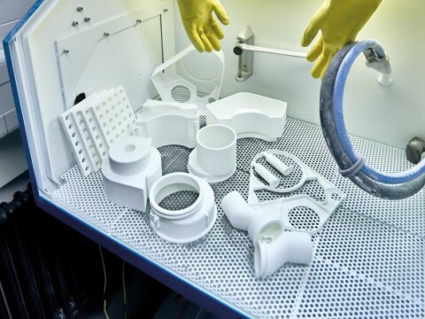 Weisse Kunststoffteile in einem Labor mit Sicherheitsausrüstung und Handschuhen, angeordnet auf einer perforierten Oberfläche.