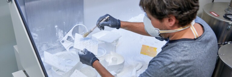 Eine Person in Handschuhen und Schutzmaske bepinselt ein 3D-gedrucktes Objekt, mit weissem Pulver umgeben.