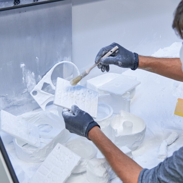 Eine Person in Handschuhen und Schutzmaske bepinselt ein 3D-gedrucktes Objekt, mit weissem Pulver umgeben.