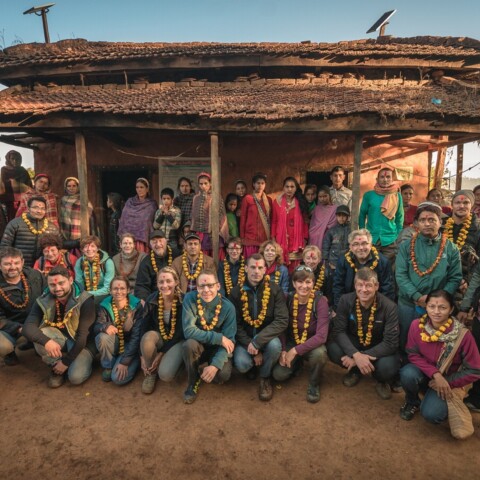 A large group of diverse people, wearing flower garlands, poses together in front of a rural building with a thatched roof.