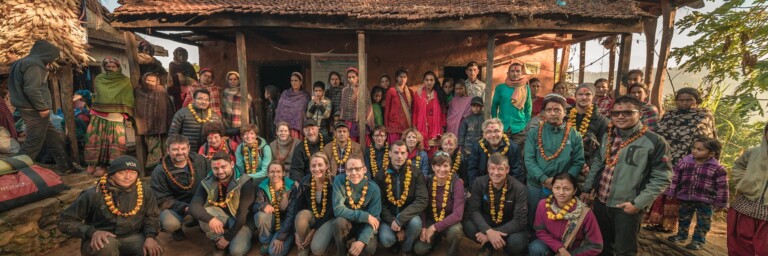A large group of diverse people, wearing flower garlands, poses together in front of a rural building with a thatched roof.