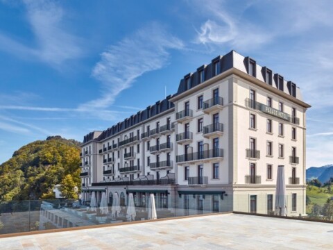 Ein modernes Hotelgebäude mit mehreren Stockwerken, grosser Fensterfront und Balkon, umgeben von Natur und blauer Himmel.