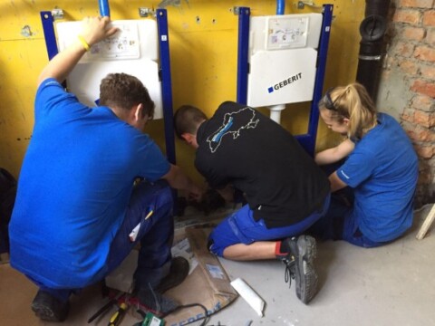 Drei Personen in blauer Arbeitskleidung montieren Sanitäranlagen an einer Wand.