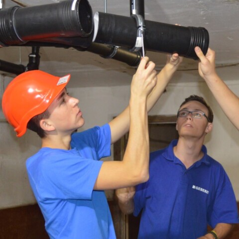 Three construction workers are assembling black pipes in a basement, wearing hard hats and safety gear.